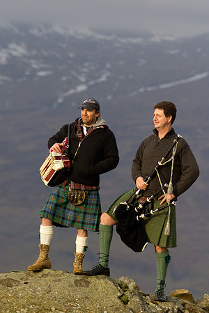 A bagpiper and accordion player holding their instruments, standing on a rock with high mountains in the background.