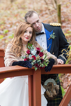 Bride and groom standing close together on a bridge, posing for a photo after their wedding ceremony. The bridge holds a large bouquet of red and white flowers.