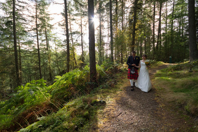 A newlywed bride and groom walk through sunny woodlands on a hill near Pitlochry, Perthshire